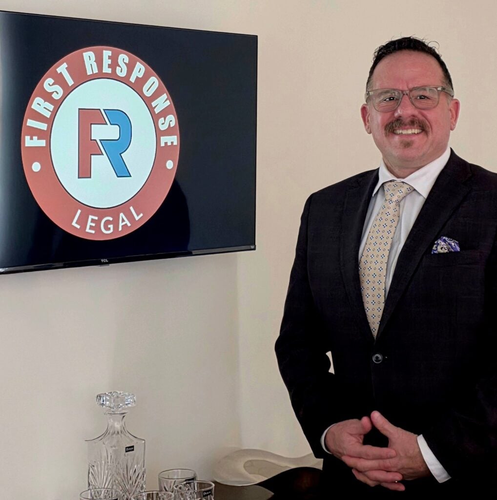Timothy Battistelli, founder of First Response Legal, standing beside a monitor displaying the firm's logo, emphasizing his commitment to serving veterans and first responders in legal matters.