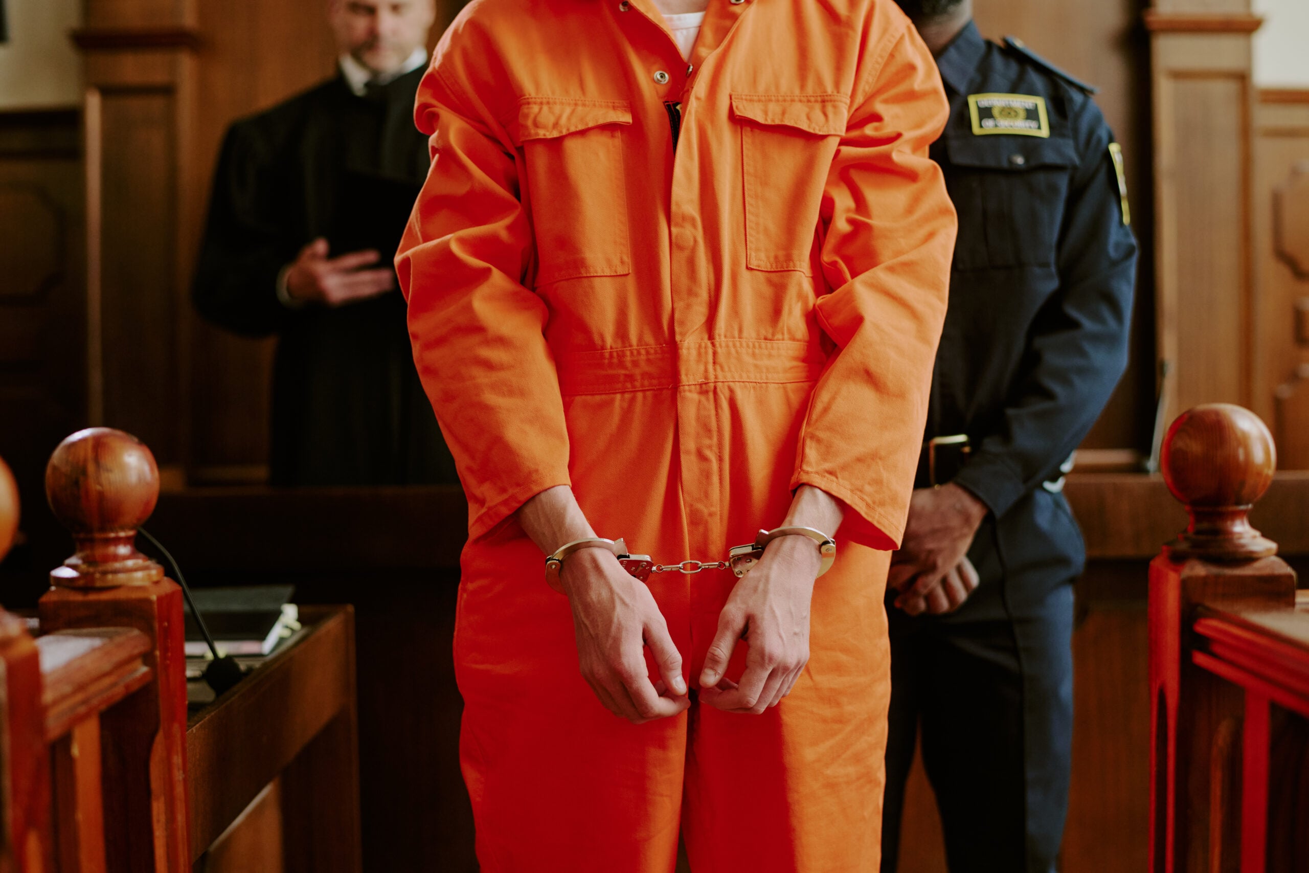 Young adult man in orange prison jumpsuit and handcuffs standing in courtroom, flanked by police officer and judge, representing felony defense context.